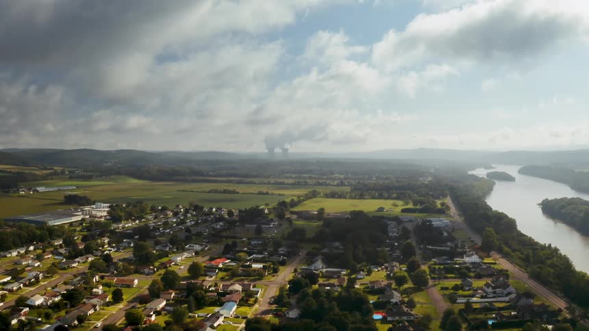 Vehicles pass through a neighborhood along the susquehanna river with steam rising from a nuclear power plant in the distance.