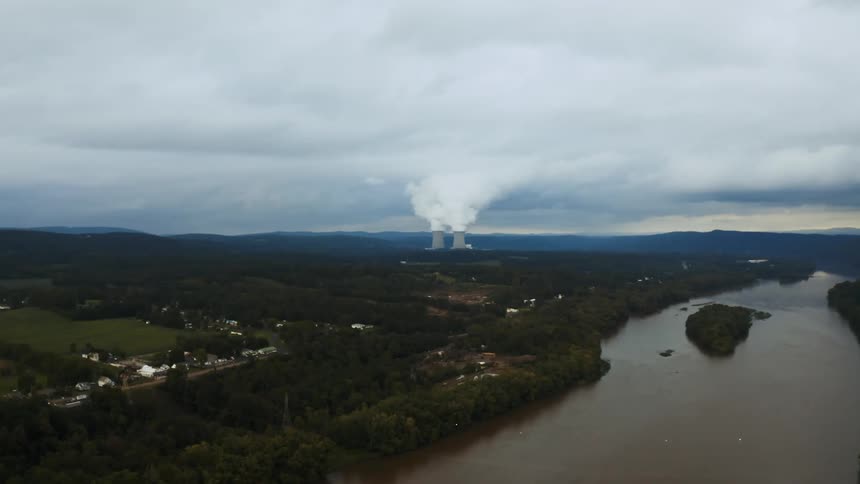 A small island sits in the middle of the susquehanna river next to a neighborhood with a nuclear power plant in the distance.