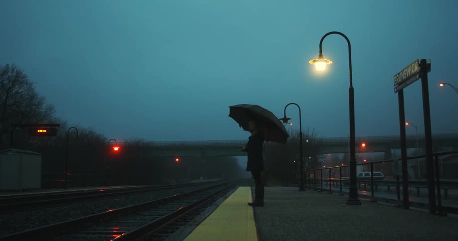 Woman with black umbrella coat and pants stands in train station as it rains