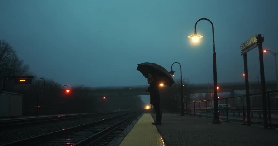 Woman with black umbrella coat and pants stands in train station as it rains at dusk