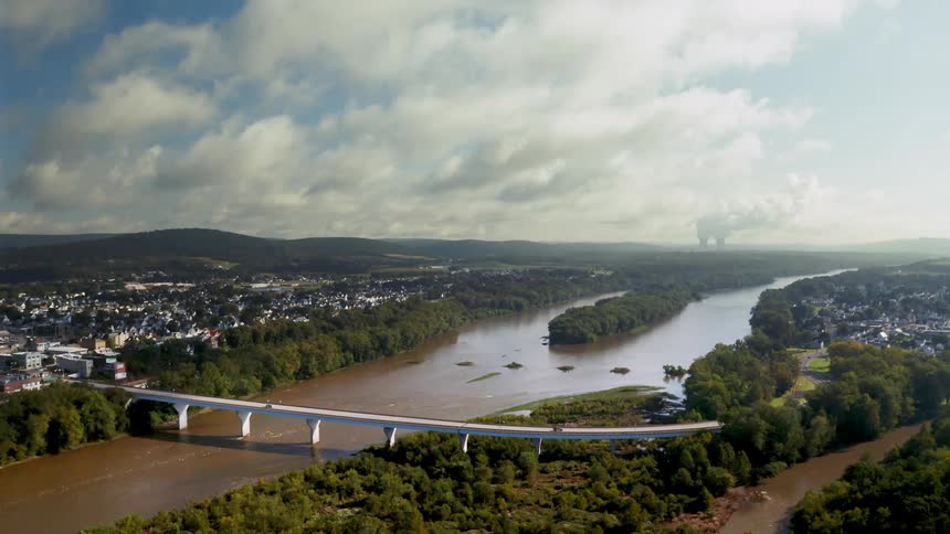 Vehicles cross a long bridge over the Susquehanna river. Steam rises from a nuclear power plant in the distance