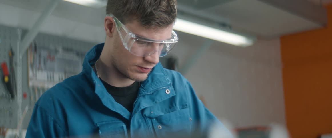 A man wearing goggles and a utility suit sits down at a table in a lab