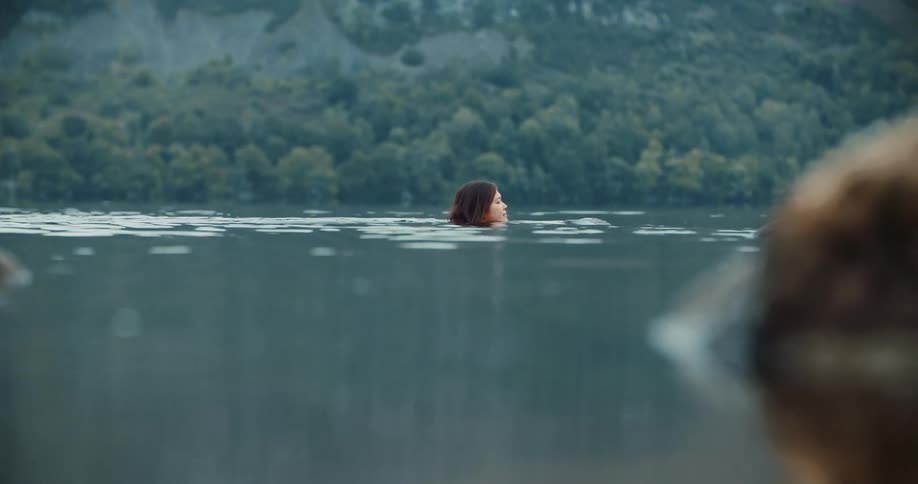 Woman with red hair swims in lake next to mountain with forest