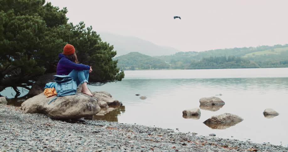 Woman with woolly hat purple jacket and jeans sits on rock watching lake and flying bird