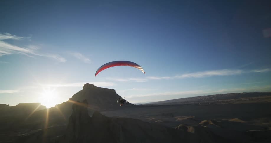 Person para glides over badlands toward sun cresting plateau topped mountain