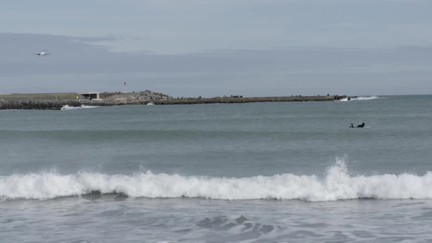 A plane flies in a cloudy sky over this beach as a surfer floats in the water