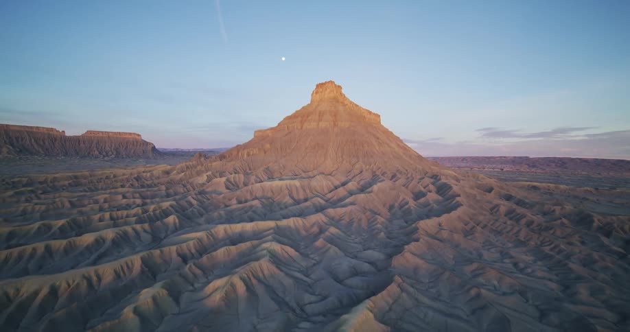 Moon sits in sky over mesa and plateaus in badlands backed by a red tinted horizon