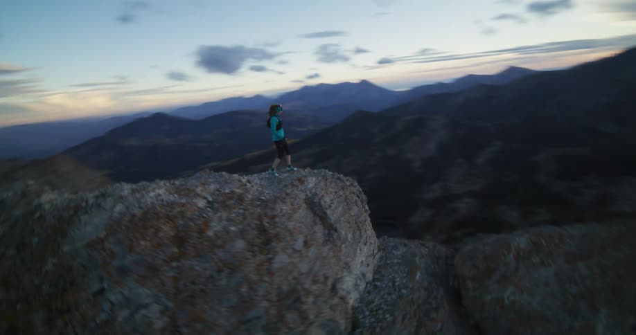 A woman wearing shorts is running along the top of a mountain ridge and she stops at the edge of a cliff