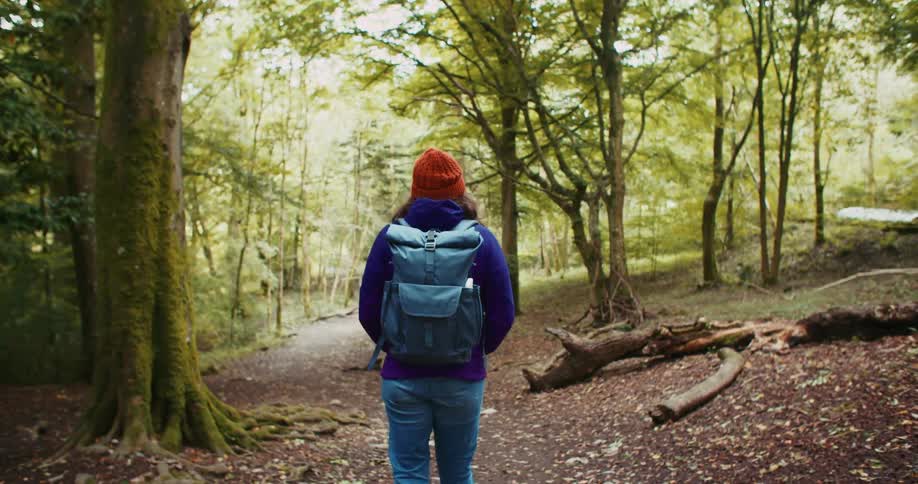 A woman wearing a backpack is walking on a trail through a forest