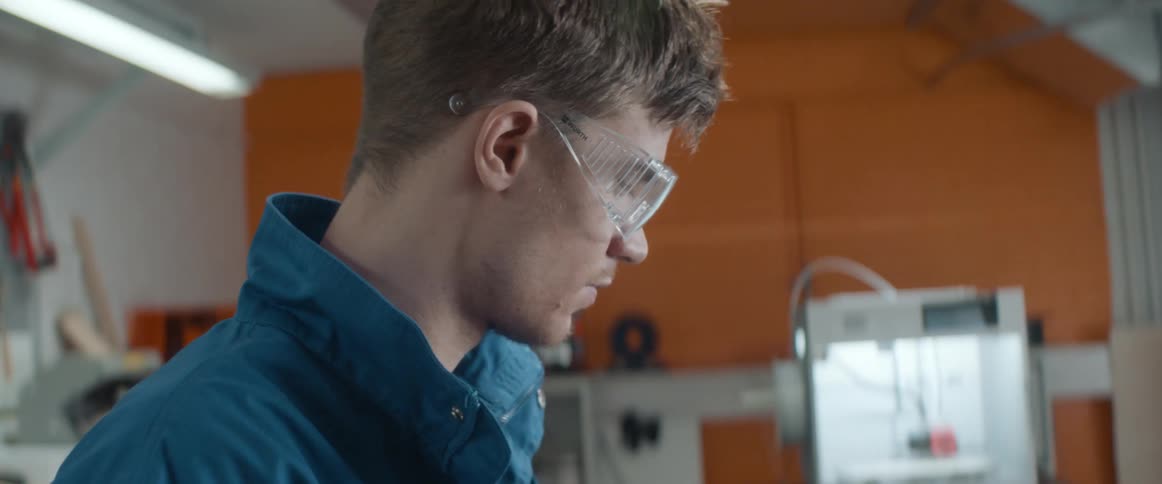A man wearing goggles sits down at a workbench to being work