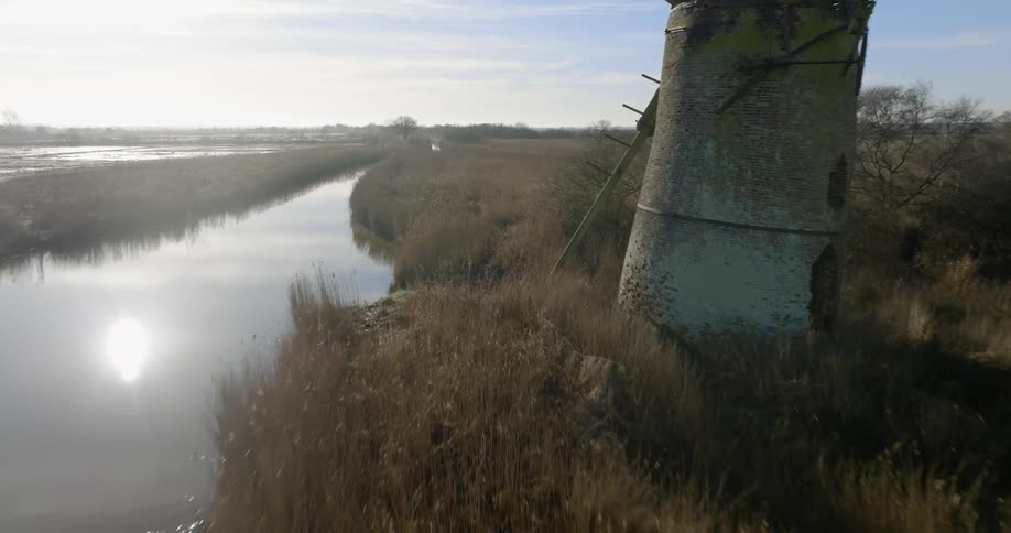 A broken down windmill stands near the river whose smooth surface reflects the bright sun