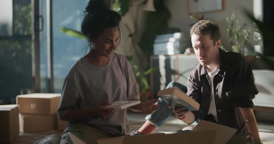 Happy young couple sitting in the living room floor unpacking books from a cardboard box