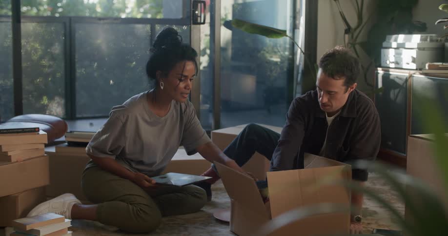 Young couple sitting in the living room surrounded by cardboard boxes checking a tablet while unpacking