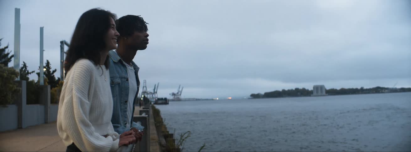 A man and woman smile as they lean against the railing and look out at the ocean on a cloudy day