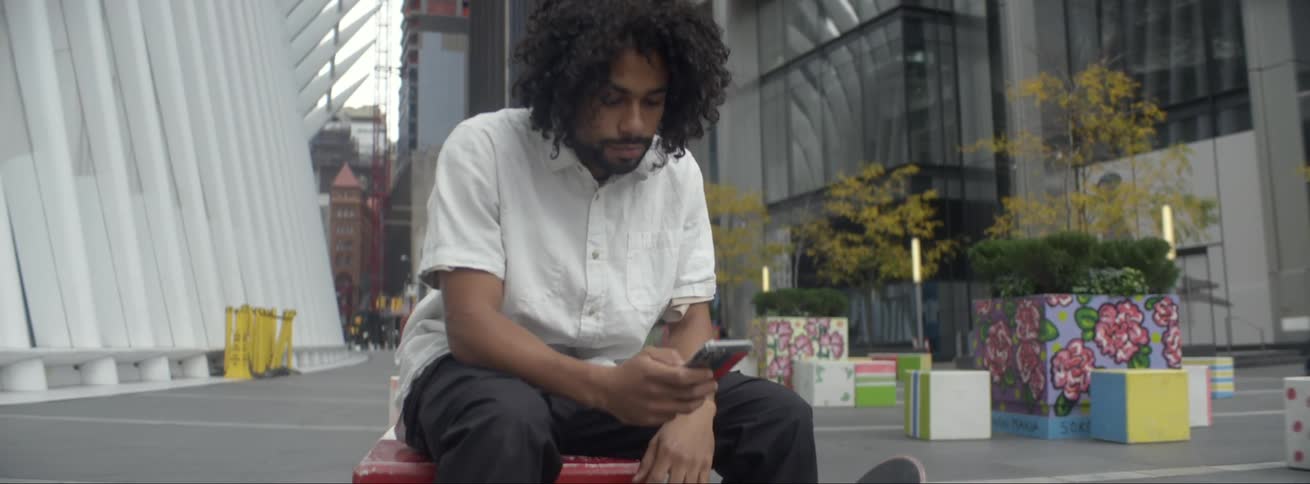 Young man sitting on a colorful bench on a plaza in the city looks at his smartphone