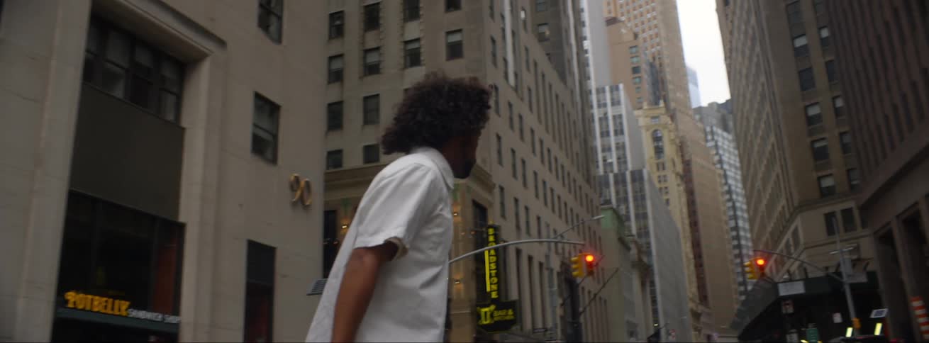 Man rides skateboard along street toward intersection passing bicycles parked cars and skyscrapers