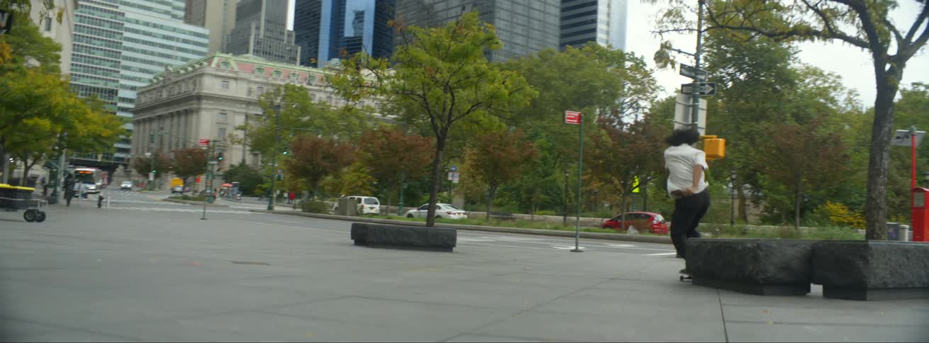 A male on a skateboard jumps over a bench in a city as pedestrians stand by and cars drive past