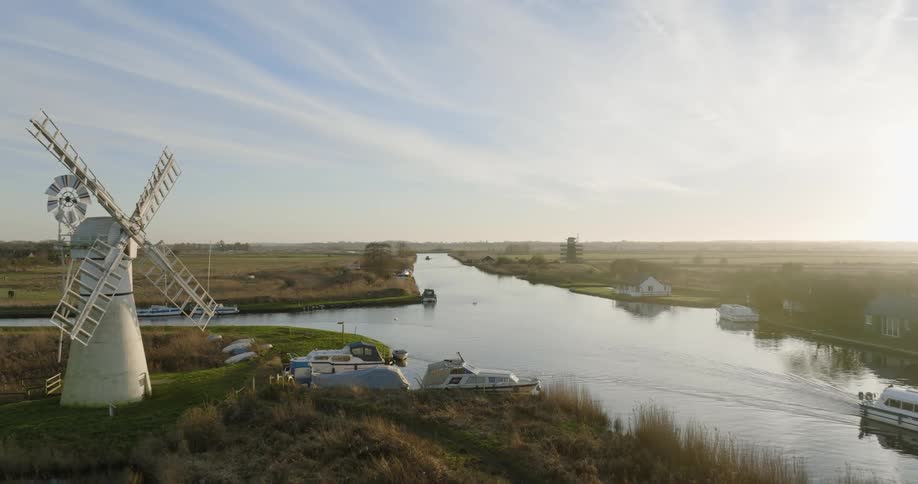 Sun brightens horizon as boat moves along river passing boats moored near windmills