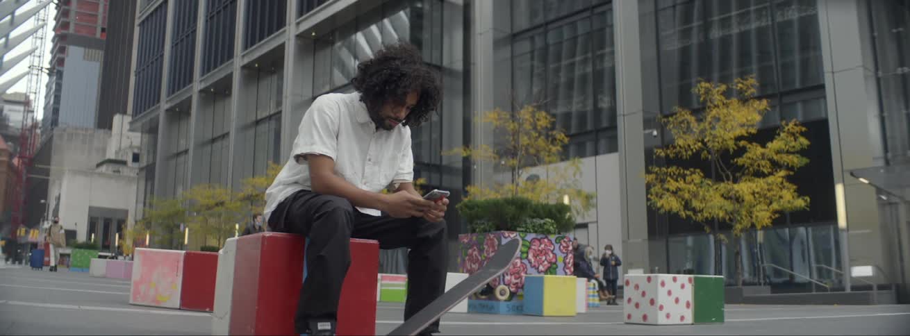 A man is sitting in a park with one foot on his skateboard as he looks at his phone