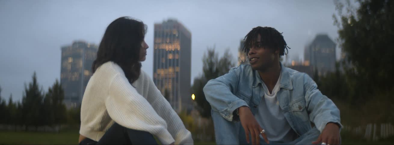 Man and woman sit outdoors in park talking with mist covered skyscrapers in the background