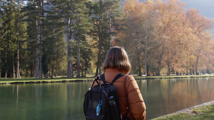 Woman wearing an orange jacket and a backpack looks at a river in a park