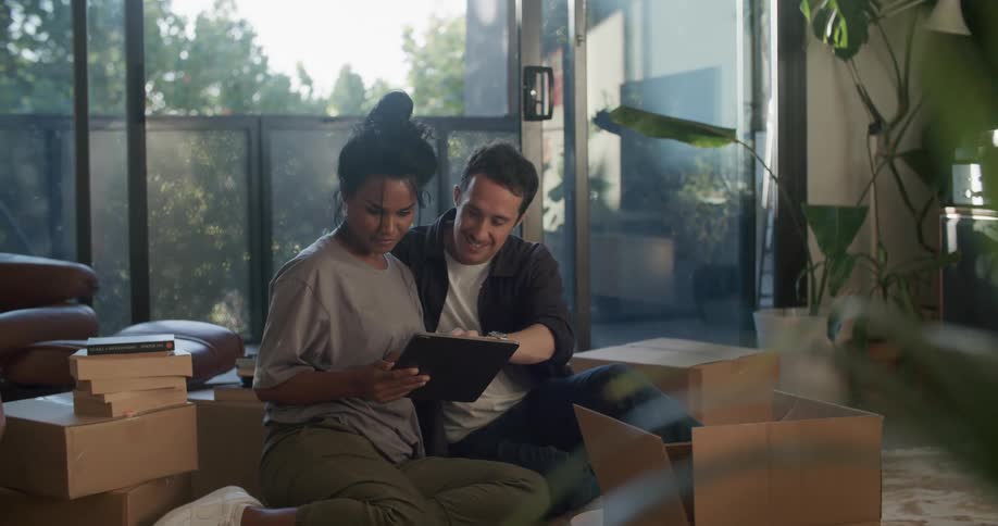 A man and woman sit around moving boxes and look at an ipad tablet together