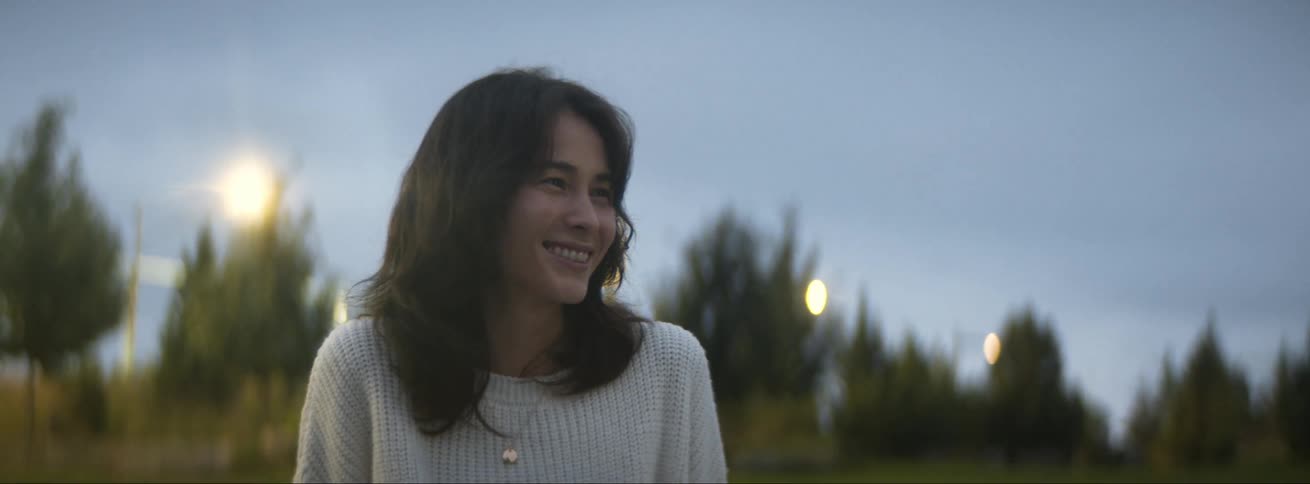 Woman with wavy hair white sweater and necklace talks and smiles at person in park at dusk
