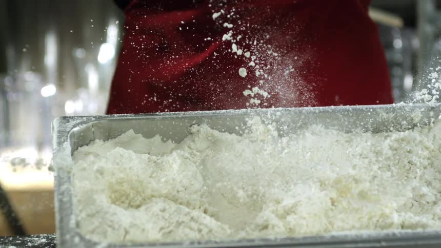 A chef wearing a red apron drops a couple of raw chicken strips inside a container full of flour