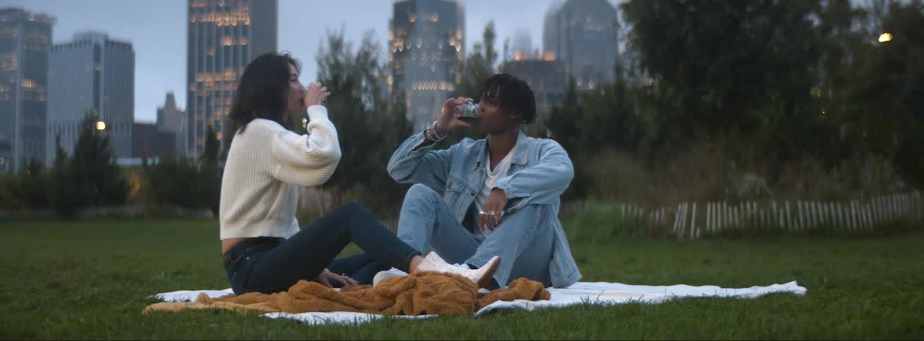 Couple sit on blanket in park drinking together with skyline in the background