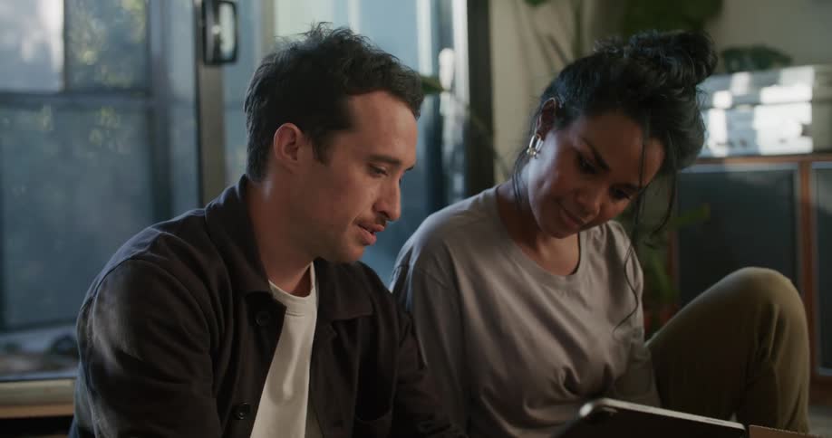 Man and woman sit on floor with stacked dishes discussing tablet contents