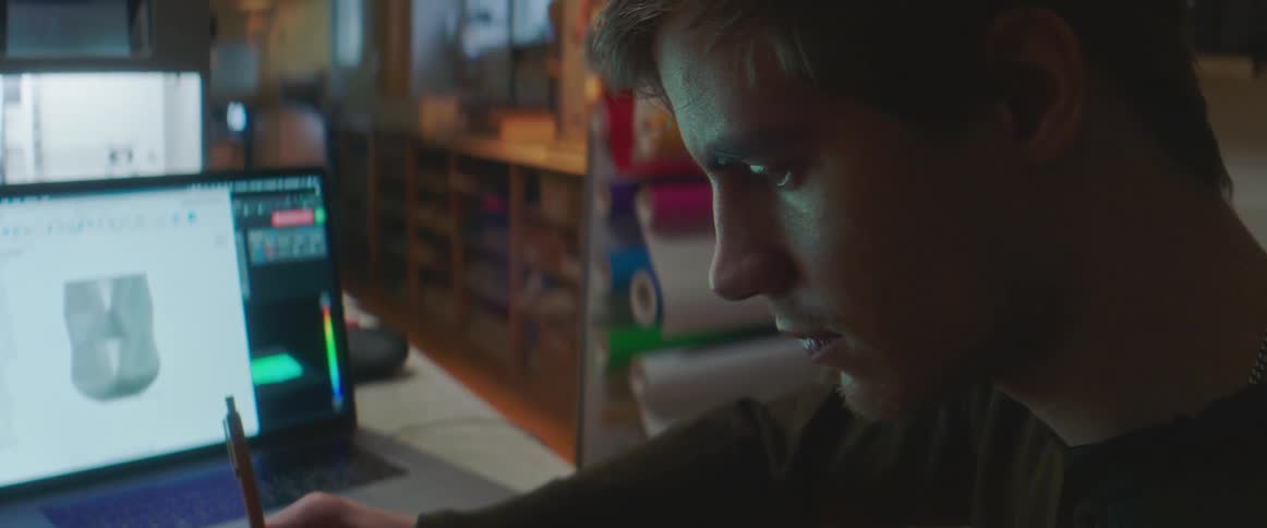 A man sits in front of a laptop with an image on it and writes on paper
