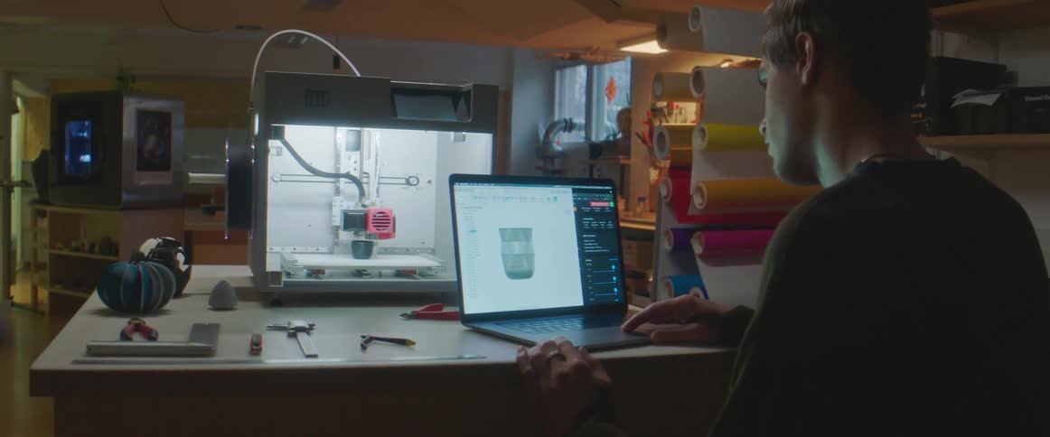 Young man working with a 3d printer using a laptop inside a dimly lit warehouse