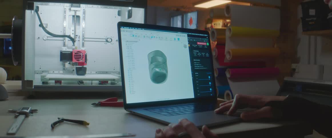 A man sits at a desk and looks at an image on a laptop while a 3d printer behind him makes the image