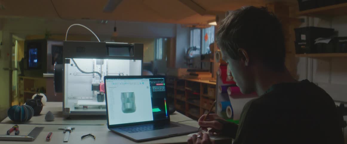 Man with black sweatshirt sitting at table works on 3d on laptop and writes down in notebook in lab