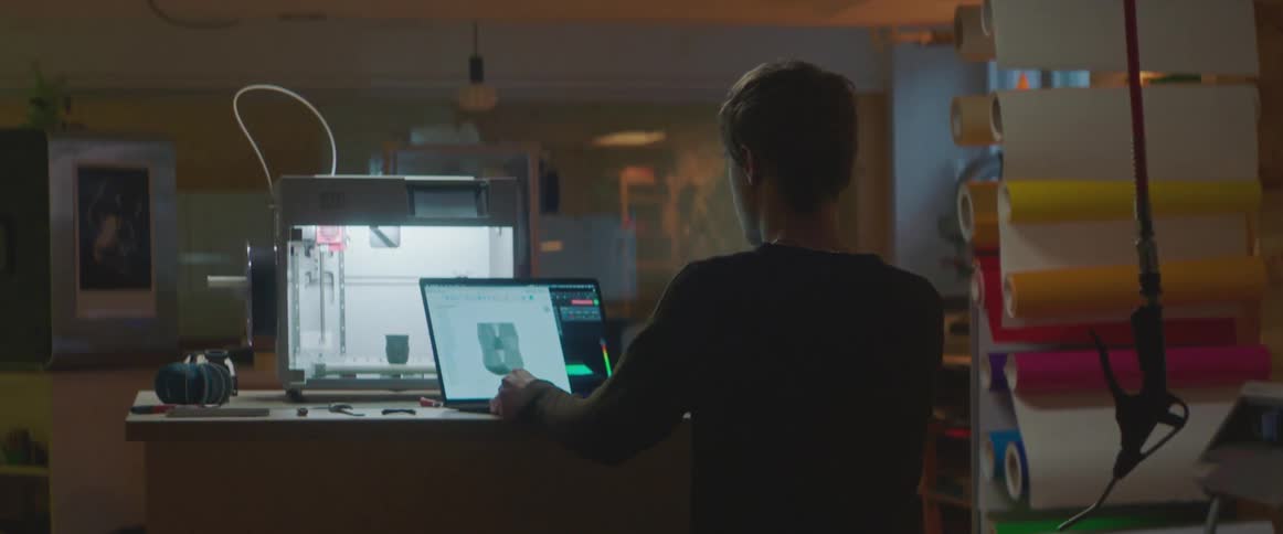 A man carries a laptop computer to a desk with a larger monitor and sits down