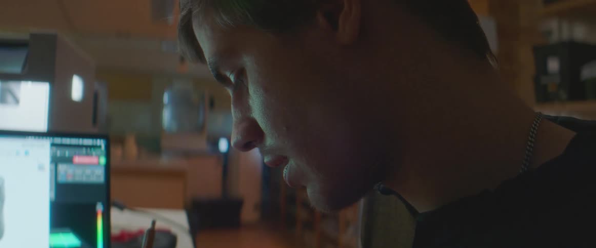A man is seated with a computer in a room filled with art supplies