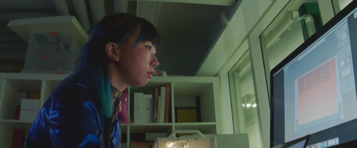 An asian woman is sitting at a desk looking at a computer screen
