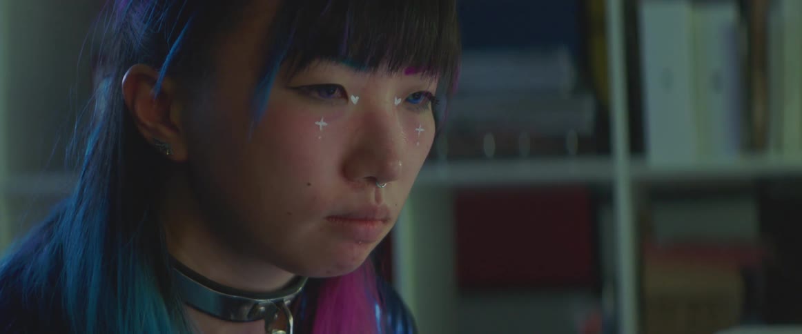 A teenager is sitting in her room and wearing colorful makeup