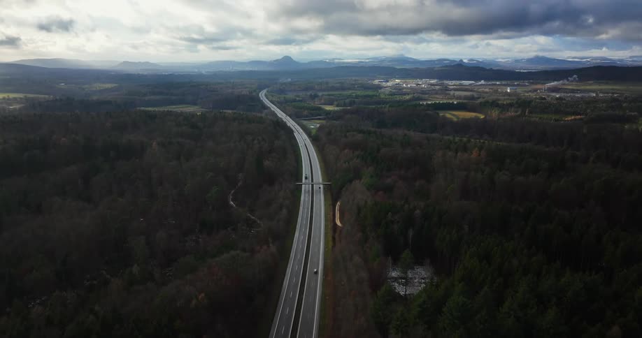Aerial view of light traffic on a long highway surrounded by forests and mountains on the horizon under a cloudy sky