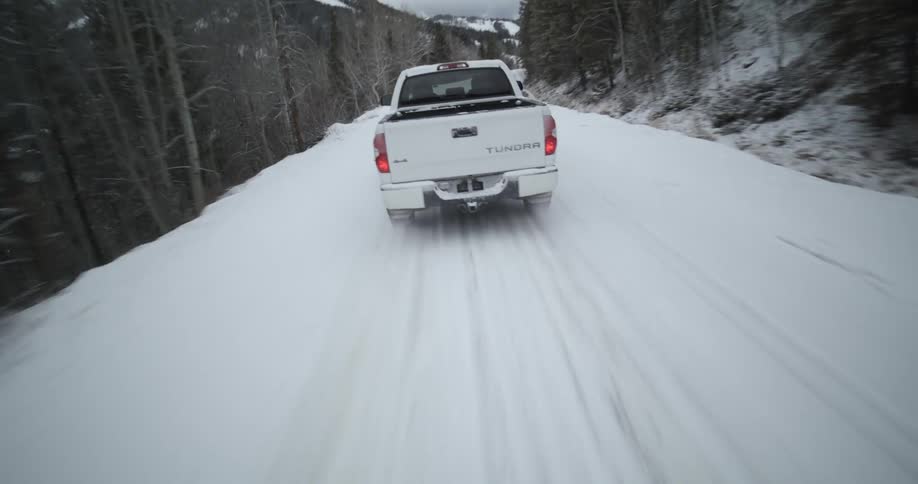 Aerial view of a white truck traveling on a snow covered forest mountain road with a mountain range in the distance