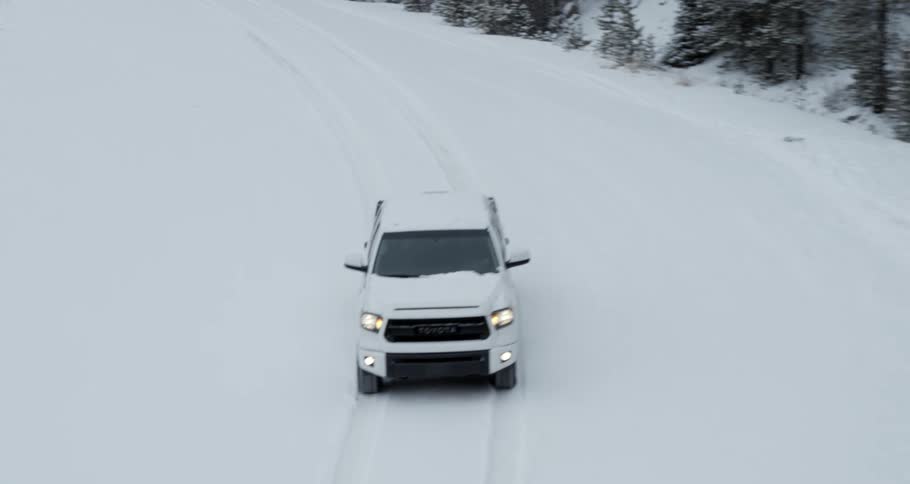 A white truck with the headlights on is traveling on a snow covered forest road
