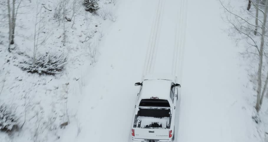 Aerial view of a white truck traveling on a snow covered forest road lined with snow covered trees