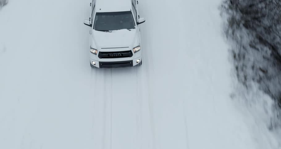 Aerial view of a white truck traveling on a snow covered forest road lined with snow covered trees