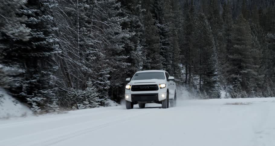 A white truck with the headlights on is traveling on a snow covered forest road