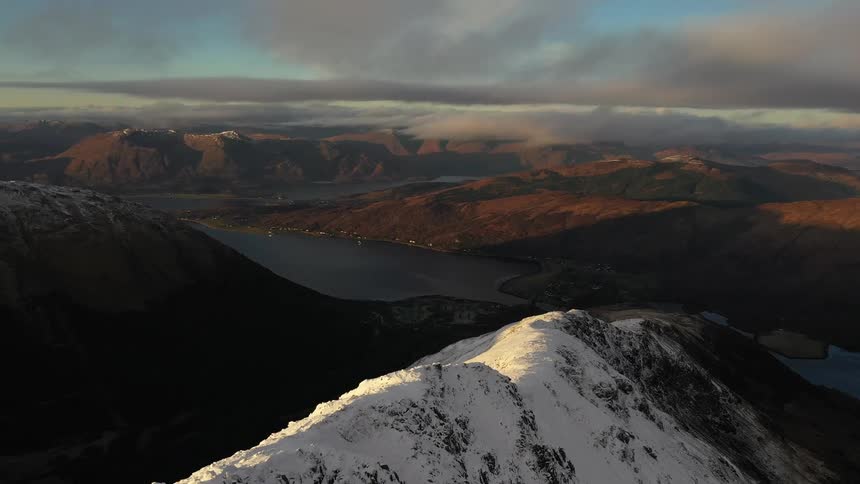 Aerial view of a snow covered mountain ridge with mountains in the distance under a cloudy sky