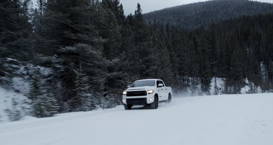 A white truck is traveling on a snow covered forest mountain road with mountains in the distance
