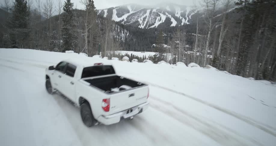 Aerial view of a white truck traveling on a snow covered forest mountain road with a mountain range in the distance