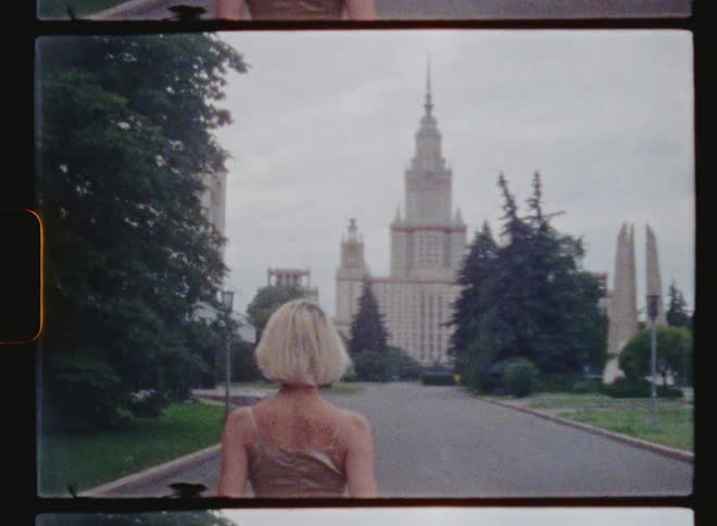 Woman walks down street toward architecturally detailed buildings before turning and posing