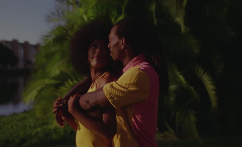 A man hugs his girlfriend from behind and kisses her on the cheek as they stand in front of a large tree