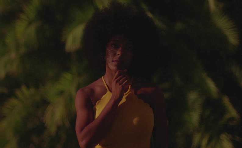 Woman with curly hair and yellow dress stares at camera touching her hair